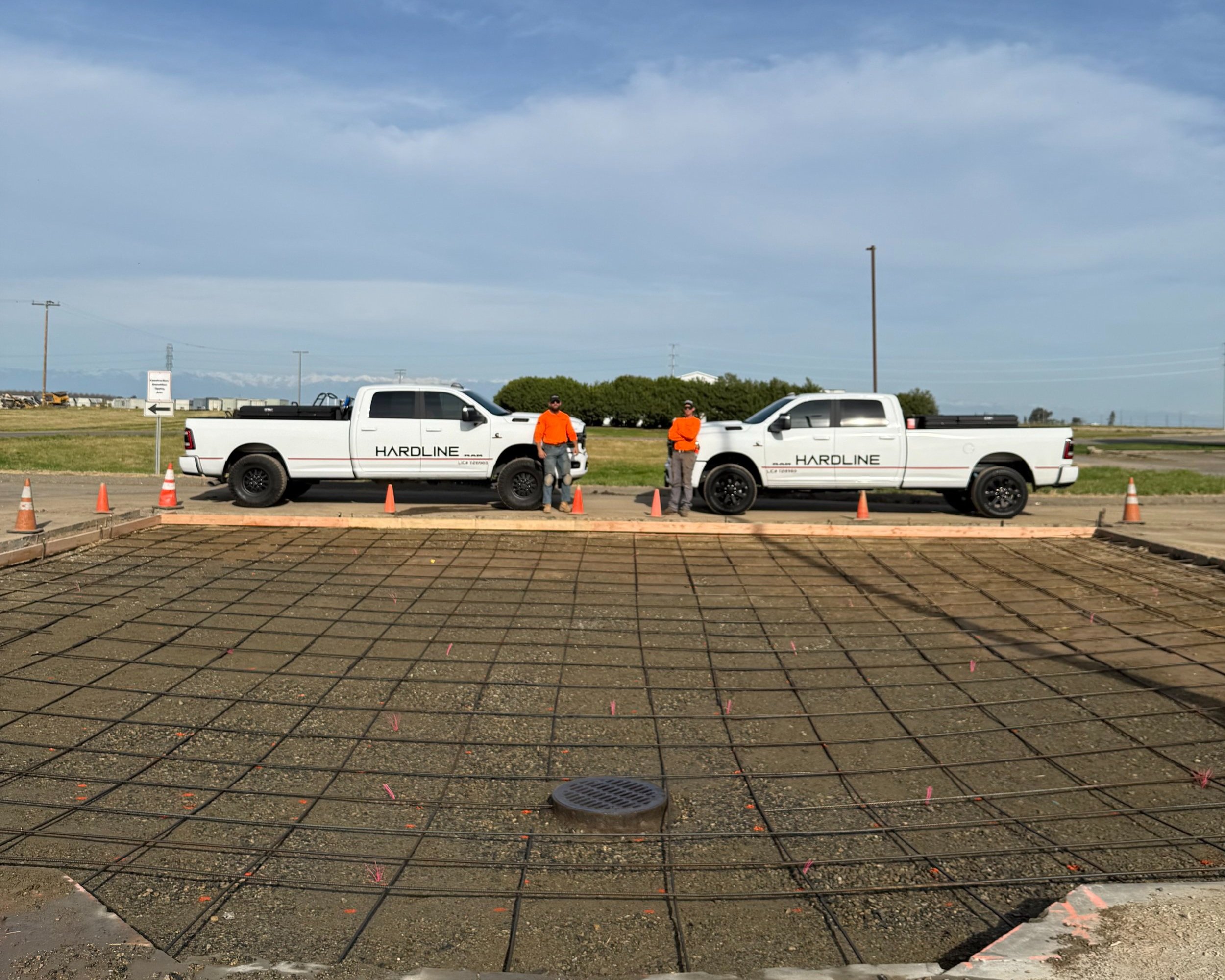 Two white pickup trucks with 'HARDLINE' branding parked on a construction site with three workers standing between them. The workers are wearing orange shirts, dark pants, and safety gear. The site has a rebar grid and building foundation in the foreground, with traffic cones surrounding the trucks.