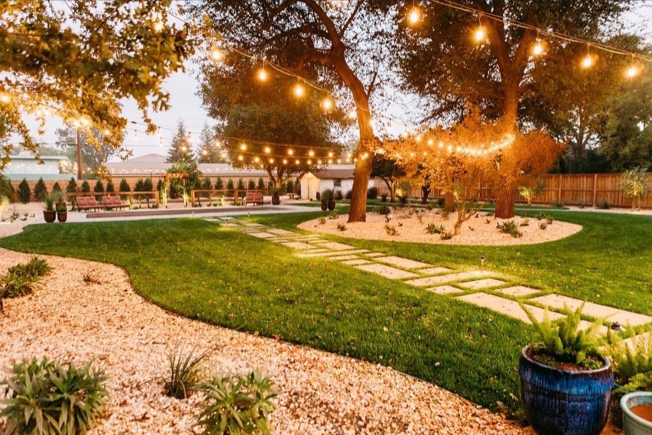A backyard garden during sunset with string lights hanging above, featuring a curved gravel pathway, lush green lawn, trees, and potted plants, enclosed by a wooden fence.